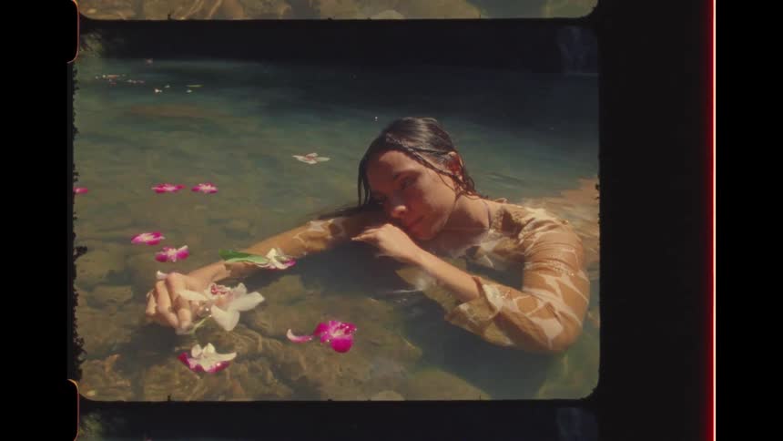 Vintage Super 8mm footage captures a serene woman in a dress floating in a sunlit natural pool amongst delicate flowers