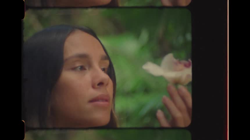 A serene woman in a tropical jungle gently touches her face with a white orchid in this vintage-style, slow-motion footage