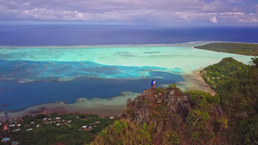 A couple embraces on a rocky mountain summit, taking in the epic aerial view of a tropical island lagoon with stunning turquoise water