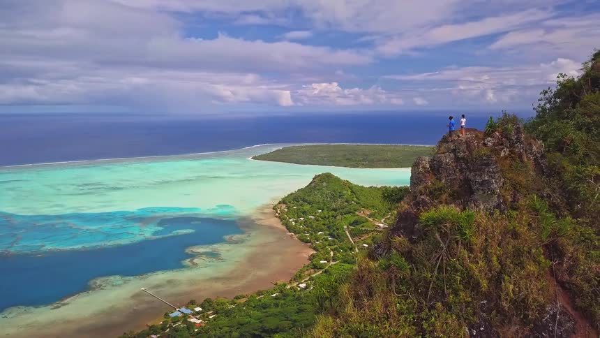 Aerial pullback reveals two hikers on a mountain summit overlooking a stunning turquoise lagoon and tropical island