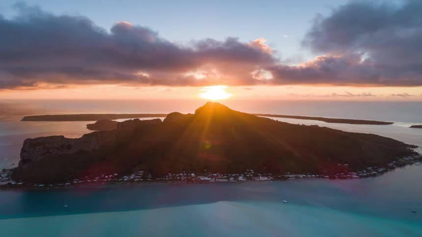 Aerial timelapse of a glorious sunset over a lush tropical island as sun rays beam from behind the mountain and clouds drift across the sky