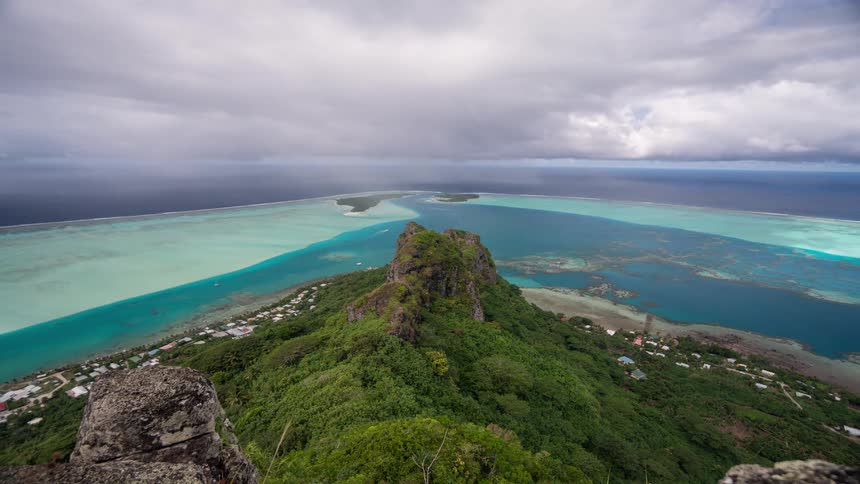 Epic time-lapse from a mountaintop vantage point shows dramatic clouds sweeping over a lush tropical island surrounded by a vibrant turquoise lagoon