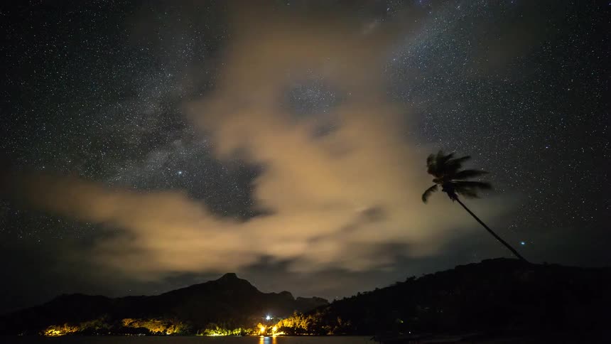 A stunning timelapse shows the Milky Way galaxy and swirling clouds moving across the night sky over a tropical island with a lone palm tree