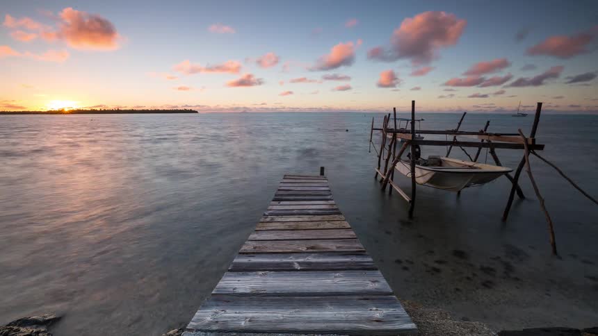 A serene time-lapse captures a golden sunrise over a tropical lagoon, viewed from a rustic wooden pier with a small boat suspended over the calm water