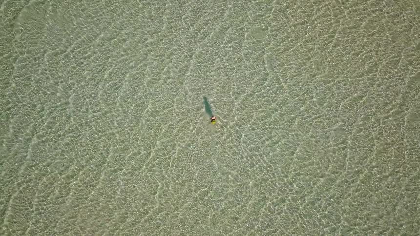 A mesmerizing top-down aerial shot captures a lone person standing in the shimmering, shallow, crystal-clear sea as the camera ascends