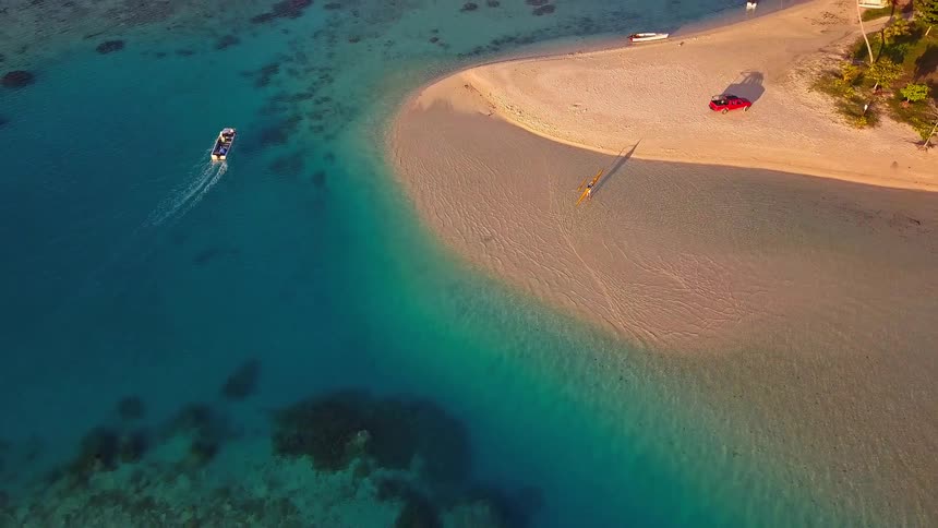 A stunning aerial view pulls back from a kayaker on a sandbar to reveal a picturesque tropical island with turquoise water and lush green mountains
