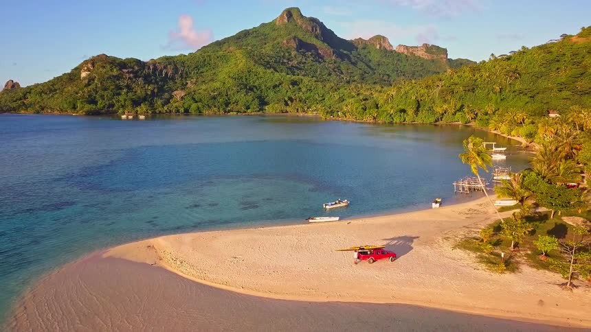 An aerial shot pulls back over a pristine tropical bay, revealing a white sand beach where a red truck with a surfboard is parked next to turquoise waters