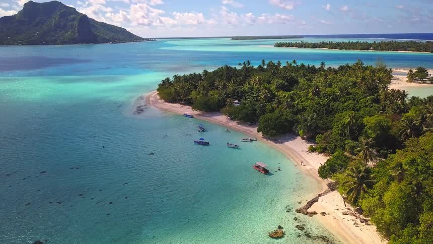 An idyllic aerial view of a tropical island in Bora Bora, with boats anchored in the shallow turquoise lagoon and lush palm trees on the white sand beach