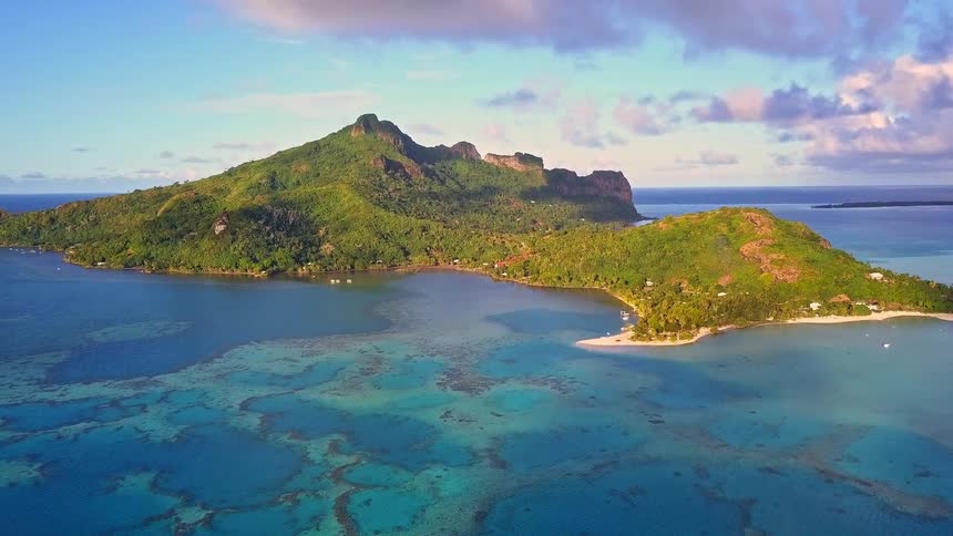 A stunning and peaceful aerial view of the lush, mountainous island of Maupiti in French Polynesia, surrounded by a vibrant turquoise lagoon and coral reef