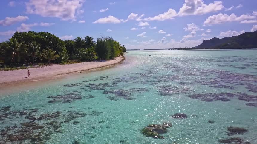 An aerial view pulls back to reveal a woman walking along a secluded white sand beach next to a vibrant turquoise lagoon in a tropical paradise