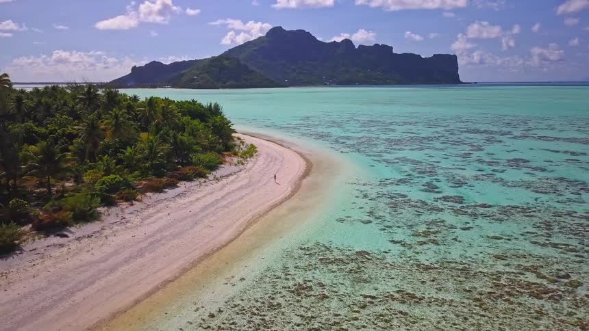An aerial drone pulls back, revealing a lone person walking on a secluded white sand beach next to a turquoise lagoon with a tropical island in the background