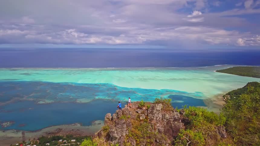 A slow aerial reveal of two hikers on a mountain summit admiring the breathtaking view of a turquoise tropical lagoon and coral reef