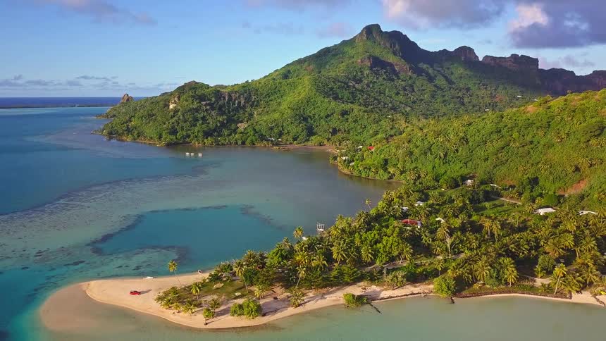 A breathtaking aerial pullback reveals a tropical paradise island with a verdant mountain peak overlooking a serene turquoise lagoon and sandy shores