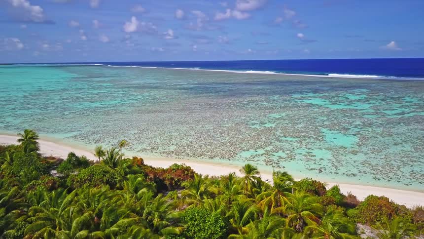 An aerial drone slowly pulls back to reveal a stunning tropical island with a white sand beach, turquoise lagoon, and lush palm trees