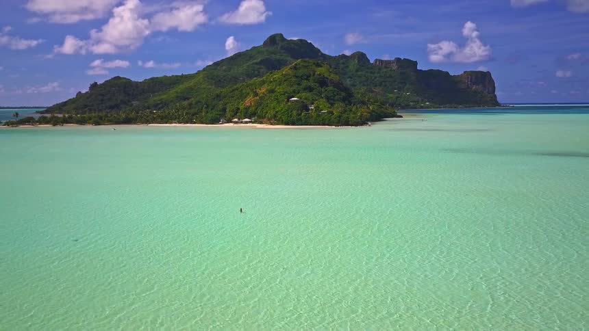 Stunning aerial footage rises above a woman wading in a crystal-clear turquoise lagoon toward a beautiful tropical island paradise