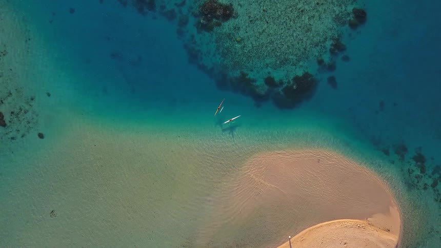 A serene aerial top-down view of two kayakers floating in stunningly clear turquoise water next to a sandy tropical island