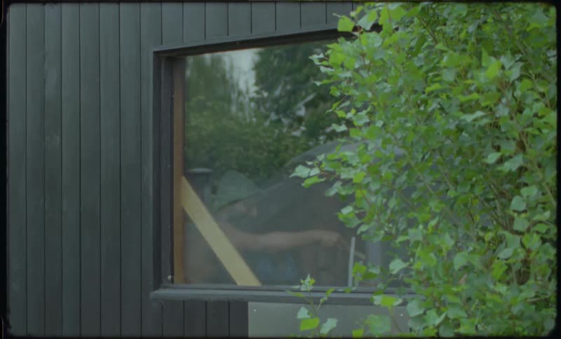 Exterior view of a man inside a modern sauna cabin, ladling water to create steam, seen through the window