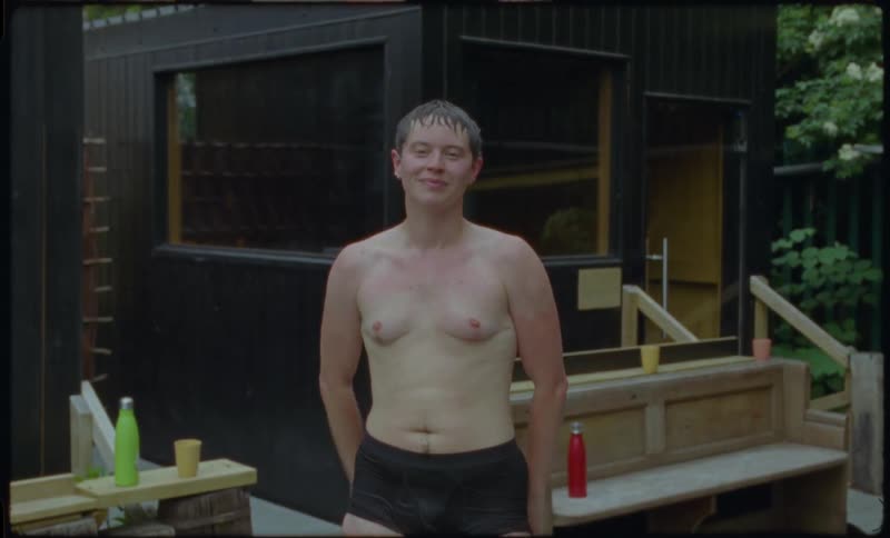 A joyful topless person with top surgery scars smiles and laughs warmly while standing outside a modern backyard sauna