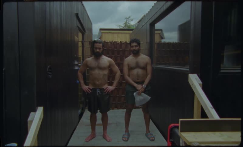 Two shirtless men, one holding a small axe, stand together between two modern buildings and smile at the camera