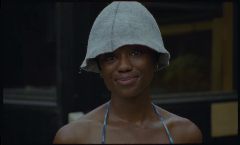 A smiling Black woman in a bikini top and sauna hat playfully looks at the camera while relaxing