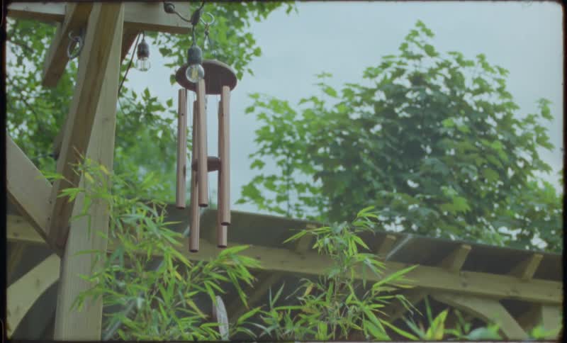 A vintage-style shot shows a peaceful wooden wind chime swaying gently from a pergola in a lush backyard garden