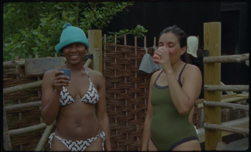 Two smiling women in bathing suits stand with drinks at a rustic outdoor sauna, enjoying a relaxing day