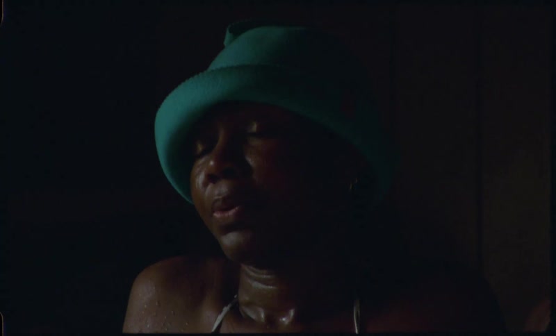 A woman in a bikini and green hat relaxes with her eyes closed in a dimly lit sauna as the camera pans down her sweaty body