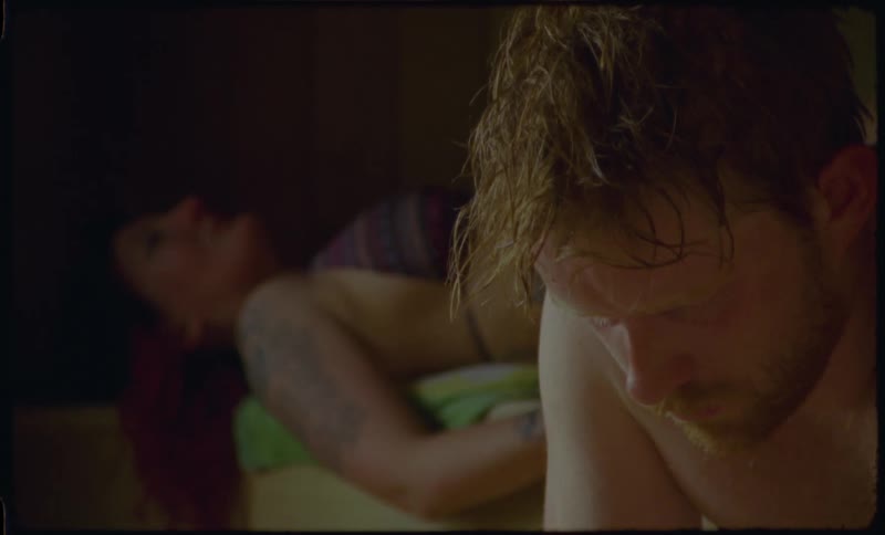 A pensive man sweats in a dimly lit sauna while a woman with red hair and tattoos rests in the background