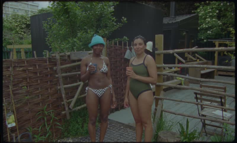 Two female friends in swimwear chat and hold drinks at a rustic outdoor sauna