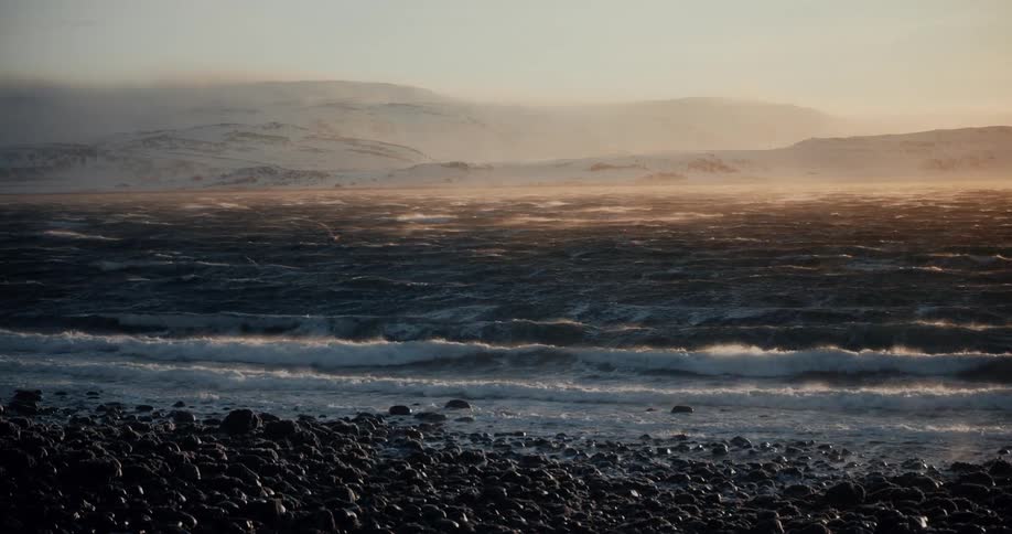 A powerful arctic wind blows over a stormy ocean against a backdrop of snow-covered mountains at sunset