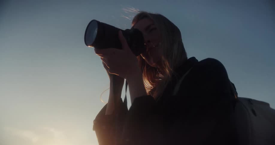 A silhouetted female photographer with windblown hair takes photos at golden hour in slow motion