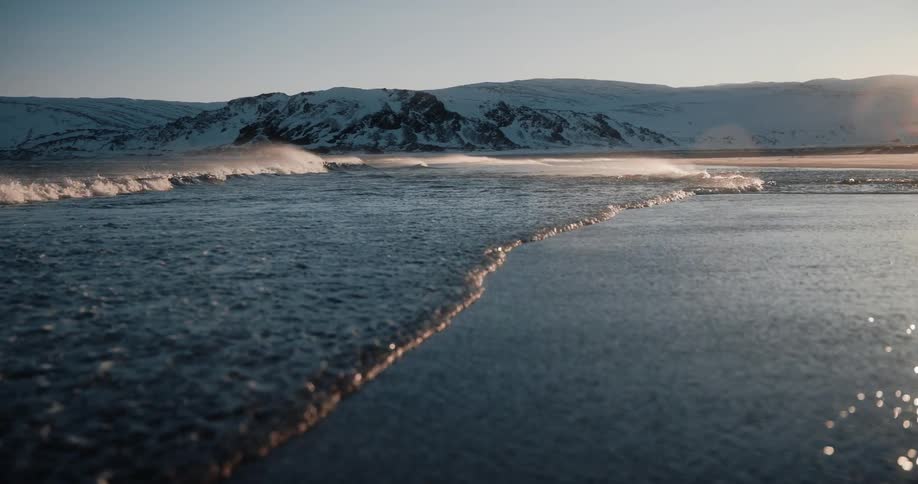 Tranquil slow-motion waves recede from a dark sand beach with a backdrop of snow-covered mountains at sunrise
