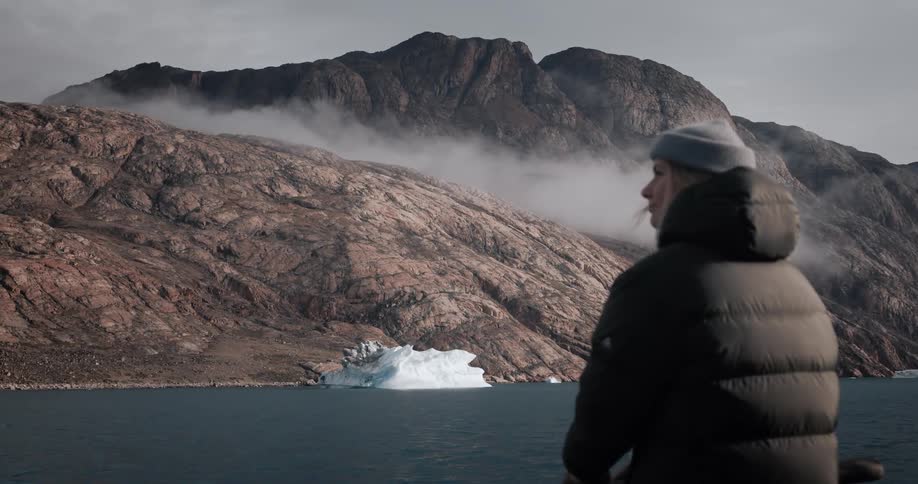 A traveler in a warm winter coat gazes out at a majestic and foggy arctic mountain landscape with an iceberg in the water