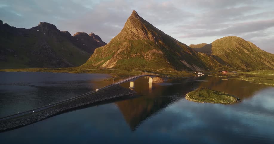 An aerial shot shows a scenic bridge over calm water with a majestic mountain reflecting perfectly at sunset in Norway