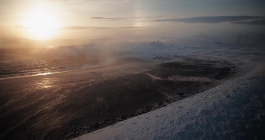 Strong winds whip snow across a dark arctic beach as the golden sun sets over the ocean