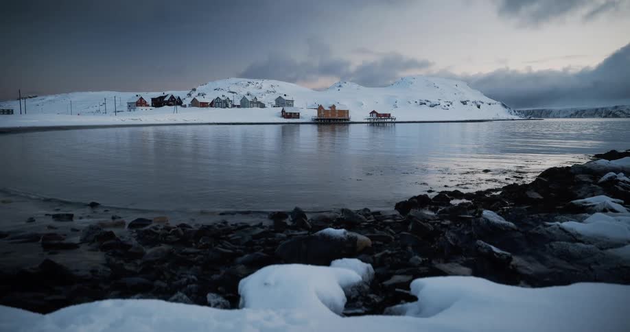 A serene view from a snowy shoreline of a remote Norwegian village nestled on a snow-covered hill across a calm bay at dusk