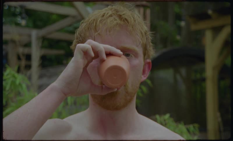 A shirtless young man with red hair drinks from a cup while standing in a lush green garden and then smiles warmly