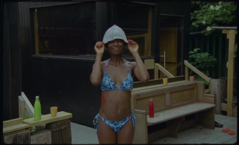 Playful young woman in a bikini and sauna hat smiles and waves at the camera outside a modern outdoor sauna, shot on vintage film