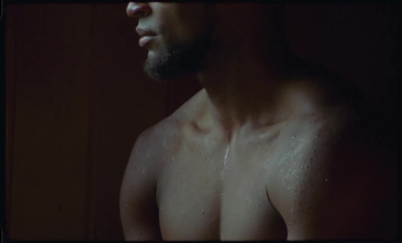 A dramatic close-up reveals the sweaty, muscular physique of a contemplative Black man in a dark room
