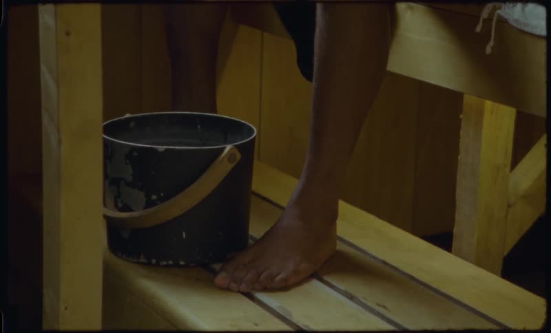 A person's bare foot rests on a wooden sauna bench as they scoop water from a bucket with a ladle