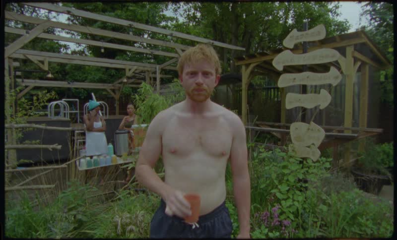 A shirtless man with red hair stands in a garden at an outdoor spa and drinks from a cup while looking directly into the camera