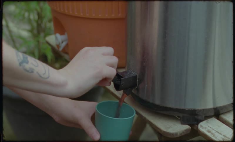 A close-up shot of a tattooed person's hands pouring a dark liquid from a large steel beverage dispenser into a green cup in a garden