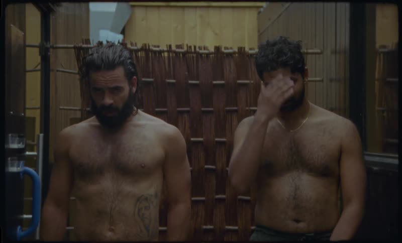 Two shirtless men sit side-by-side in a hot sauna, starting to feel the heat as they sweat and yawn in a static portrait shot