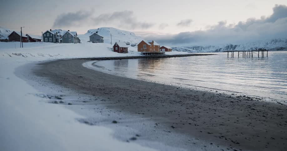 A tranquil wide shot of a snowy beach meeting the calm water with a quaint Nordic village in the background during winter