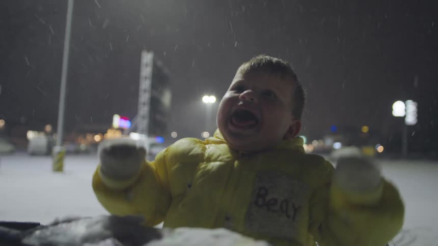 An ecstatic toddler in a yellow winter coat laughs with pure joy while catching falling snowflakes in a shopping cart at night
