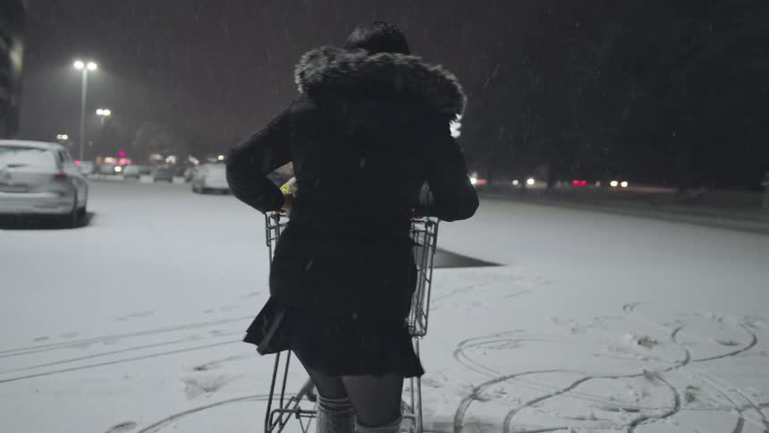A happy mother playfully pushes her laughing toddler in a shopping cart through a snowy parking lot on a winter night