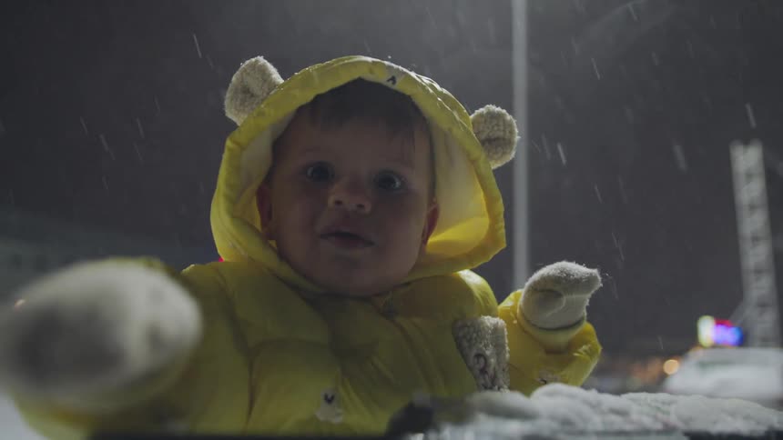 An adorable baby in a bright yellow snowsuit with bear ears watches the snow fall in slow motion with wonder on a winter night