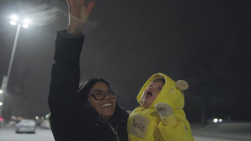 A happy mother holds her adorable baby in a bright yellow snowsuit, both looking up with joy and wonder as snow falls around them at night