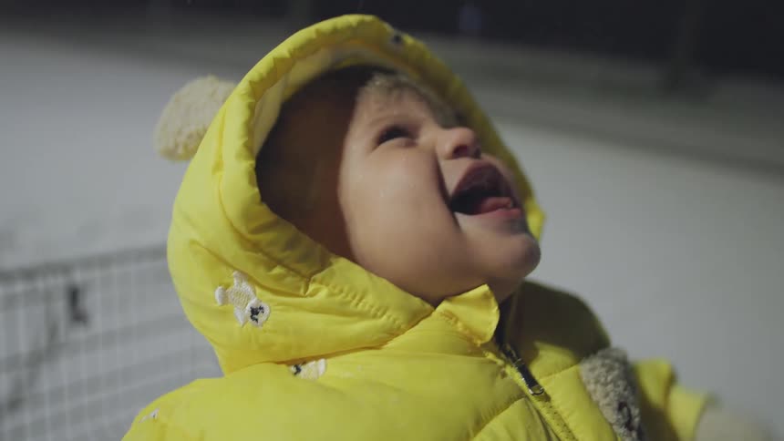 A happy toddler in a yellow hooded jacket joyfully catches falling snow on their tongue at night in slow motion