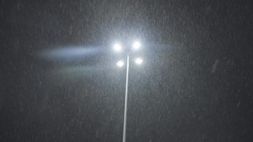 A modern street light with four bright bulbs illuminates heavy, falling snow during a dark winter night